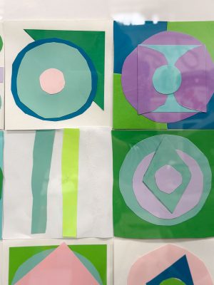 A display of colorful geometric paper art featuring circles and diamonds in shades of blue, pink, purple, and green, mounted in a grid on a white background.