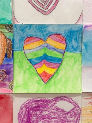 A colorful drawing of a heart with rainbow stripes set against a blue and green background. The heart is outlined with glitter, adding a sparkly effect. The artwork is part of a collection displayed on a wall.