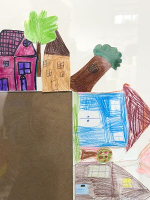 Children's drawing featuring colorful houses, trees, and geometric shapes. A red house with a purple roof, a brown house, and a blue house are visible, along with green trees and abstract forms on a light background.