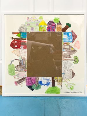 Framed artwork with colorful drawings of various houses surrounding a central blank brown area. Each house is uniquely designed with different shapes and colors, creating a whimsical border around the empty center.