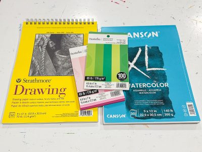 A collection of art supplies: a yellow Strathmore drawing pad, Canson watercolor paper pad, and two packs of colorful Reston Lloyd index cards. The items are on a white surface.