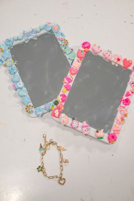 Two rectangular mirrors are decorated with colorful, whimsical designs, including hearts, stars, and animals. A gold chain with various pendants, such as a heart and a star, is placed below the mirrors on a light surface.