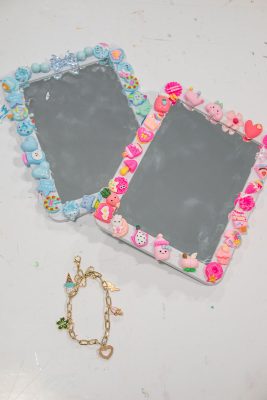 Two rectangular mirrors bordered with colorful, playful clay decorations, including ice creams, fruits, and hearts. One frame is blue-themed, the other pink. Below the mirrors lies a gold bracelet with various charms. They rest on a white surface.