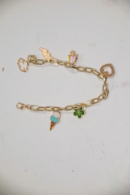 Gold charm bracelet with various charms including a cloud, lightning bolt, ice cream cone, beverage cup, clover, and a heart. The bracelet is laid out on a light, plain surface.