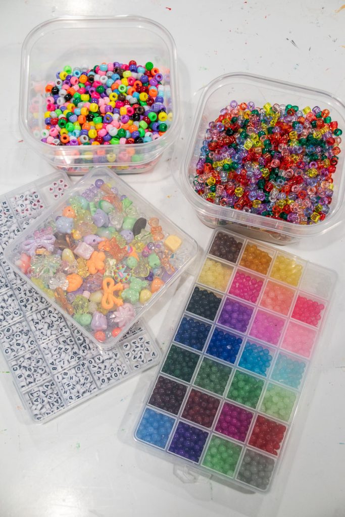 An assortment of colorful beads in various containers. There are small translucent beads, larger multicolored beads, alphabet beads, and a grid organizer with beads in a rainbow of colors, all arranged on a white surface.