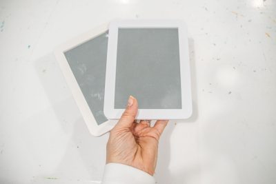 A hand holding two white tablet-like devices overlapping on a white surface. The devices have black screens with visible smudges, and the hand is wearing a white sleeve.