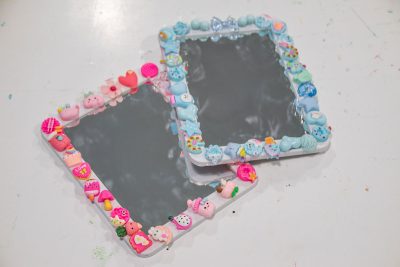Two decorative mirrors with colorful clay charms around their borders. One mirror features charms in pinks and reds, while the other has blues and pastels. The charms resemble various cute objects like animals and sweets. The mirrors rest on a white surface.