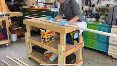 A person wearing gloves works on a long white PVC pipe at a wooden workbench in a workshop, surrounded by tools, boxes, and equipment.