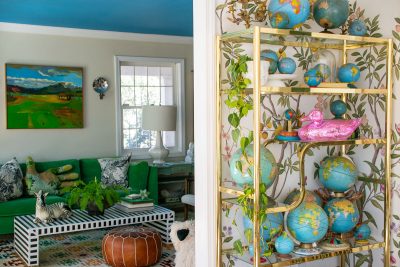 A vibrant room features a green sofa, striped coffee table, and a brown pouf. A shelf displays various globes and a pink duck sculpture. The walls have leafy patterns, and a colorful painting hangs by the window. A plant adds greenery to the room.