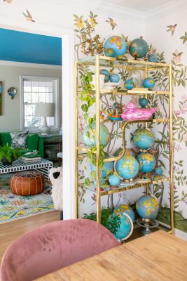 A stylish room featuring a gold shelf filled with various globes, set against a wall with floral and bird wallpaper. A glimpse into a vibrant living area with a green sofa and patterned rug. A pink chair is partially visible in the foreground.