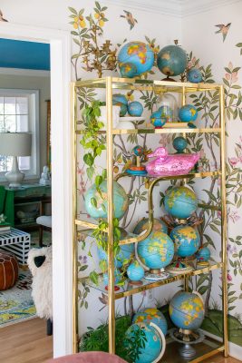 A gold shelf filled with various globes and a pink decorative object. The wall behind features floral wallpaper with birds. A leafy plant trails down the shelf. A glimpse of a colorful room is visible through the doorway.