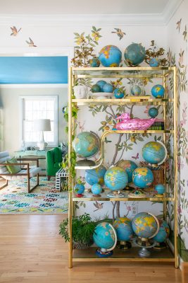 A colorful room with a large, gold shelving unit displaying various globes of different sizes. The walls are adorned with floral and bird designs. A bright, eclectic living space is partially visible in the background, featuring a green sofa.
