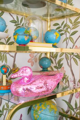 A shelving unit displaying globes and a pink, sequined duck figure under golden shelves against a nature-themed wallpaper with branches and leaves. Globes vary in size and styles, adding a vibrant, eclectic touch to the decor.