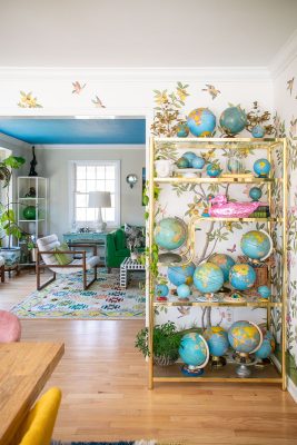 A colorful room with a shelf holding various globes and plants. The walls are decorated with bird and leaf motifs. In the background, a room with blue ceiling, a rug with geometric patterns, and green furniture is visible.