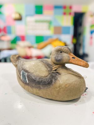 A realistic duck decoy sits on a white surface in a colorful room. The background features a blurred, vibrant patchwork of pastel blues, pinks, greens, and yellows, with a gold lamp and various blurred objects in the distance.