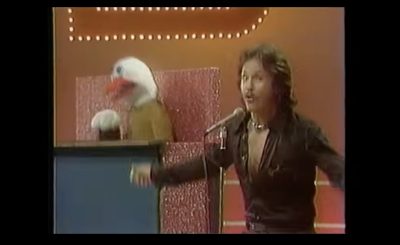 A person energetically performs on stage at a microphone, wearing a dark shirt. In the background, a large puppet resembling a bird with a white head and red beak is visible behind a podium. The stage is lit with decorative lights.