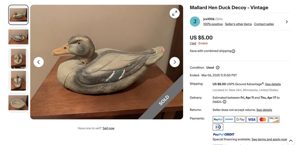 A wooden mallard hen duck decoy is displayed on a wooden surface. The decoy has detailed painting to resemble a duck, with a realistic color scheme. A "SOLD" banner is across the image. The listing shows a $5 price with shipping details.