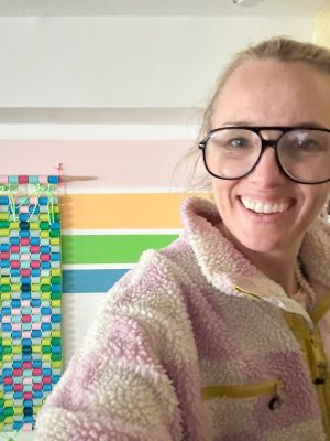 A smiling person wearing large glasses and a fluffy striped jacket stands in front of a colorful wall and a woven, multicolored hanging decoration.