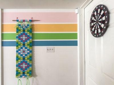 A colorful wall hanging made of interwoven paper loops hangs on a pastel-striped wall next to a white door with a dartboard mounted on it. A light switch is visible beneath the wall hanging.