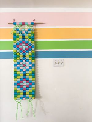 A colorful woven wall hanging made of interlaced paper strips in blue, green, and pink is displayed on a wall with horizontal pastel stripes in pink, yellow, green, and blue. A light switch is visible nearby.