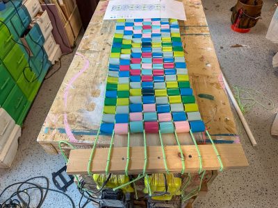 A colorful glass weaving project in progress sits on a workbench, with bright rectangular glass pieces woven together using green string. A pattern guide and various workshop tools are visible nearby.