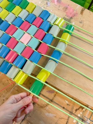 A hand weaving neon green rope through rows of colorful oval beads, arranged in a pattern on a wooden surface. The beads are blue, green, gray, yellow, pink, and red.