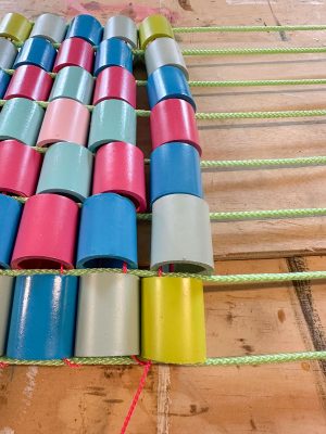 Colorful, cylindrical wooden beads are threaded onto green cords, creating a grid-like pattern on a wooden surface. The beads are painted in various shades of blue, pink, green, red, and gray.