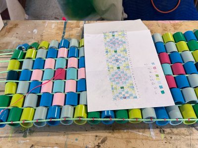 A weaving project on a wooden table uses colorful plastic cups threaded with cords, next to a sheet of paper with a color-coded grid pattern and handwritten notes.