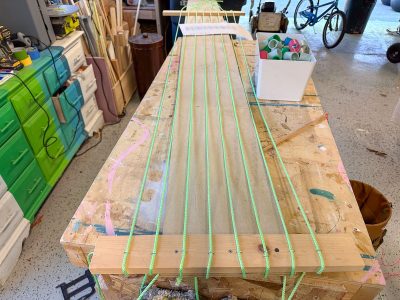 A wooden board on a workbench has green string stretched and tied across it in parallel lines. Workshop tools and materials are visible in the background.