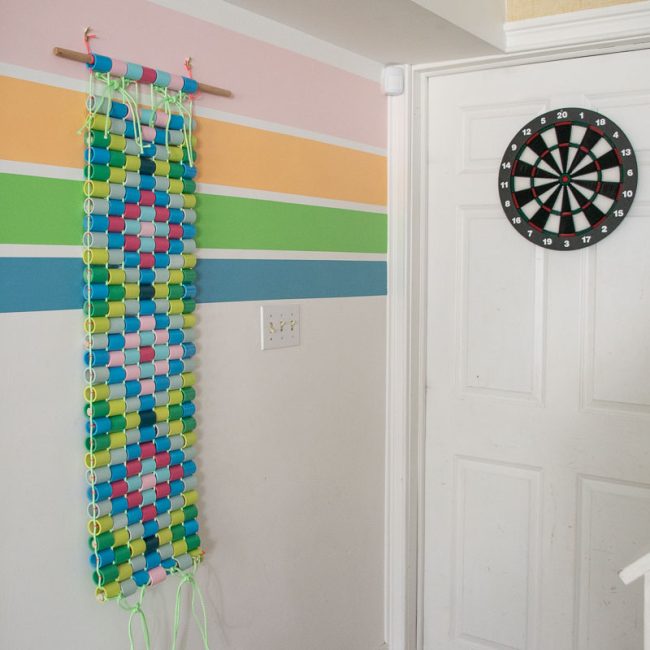 A colorful paper chain decoration hangs on a pastel-striped wall next to a white door with a dartboard mounted on it. The scene is bright and playful, with a staircase visible in the bottom right corner.