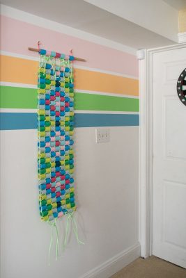 A colorful paper chain wall hanging, woven with blue, pink, green, and yellow loops, is attached to a dowel and hangs from a pastel-striped wall next to a white door.