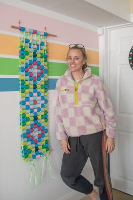 A smiling woman stands indoors beside a colorful paper chain wall hanging on a wooden dowel. The wall behind her has horizontal pastel stripes. She is wearing a checkered fleece jacket and dark pants.