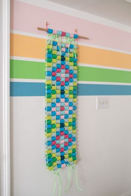 Colorful paper chains woven into a rectangular wall hanging, suspended from a wooden dowel, displayed on a wall with pastel horizontal stripes.
