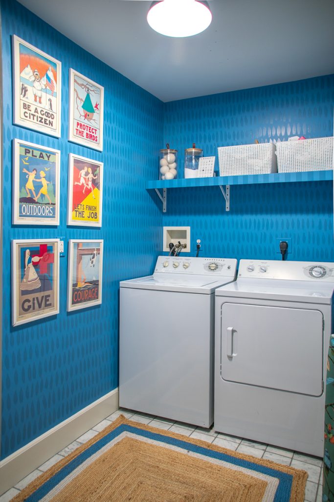 A laundry room with blue patterned wallpaper, white washer and dryer, and six vintage-style posters framed on the wall.