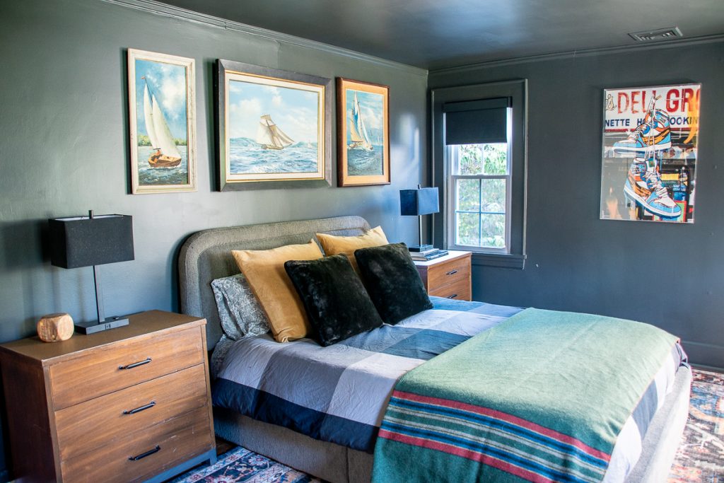 A bedroom with dark gray walls features a bed with gold and black pillows, a striped blanket, and three framed paintings of sailboats above the headboard, with a framed sneaker poster on the adjacent wall.