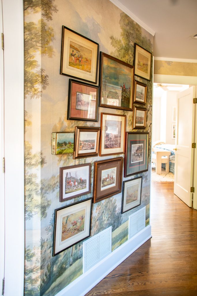 A gallery wall of various framed hunting and landscape prints arranged on a wall covered in scenic mural wallpaper.