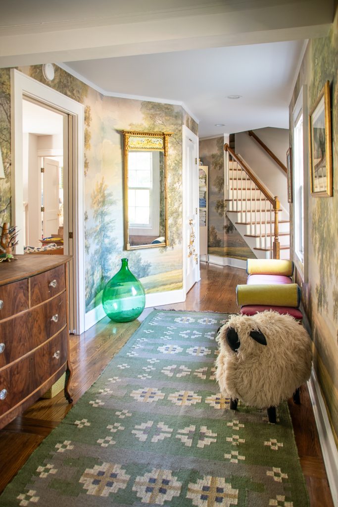 A hallway featuring a landscape mural, a green glass floor vase, a framed mirror, a patterned runner rug, and a sheep-shaped footstool.