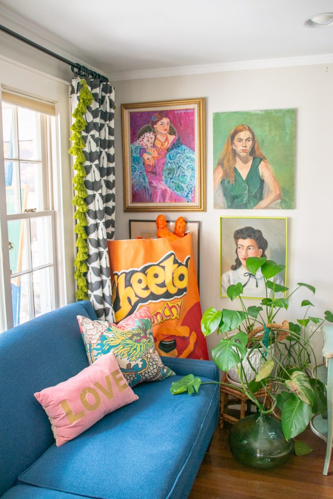 A blue sofa with a pink “LOVE” pillow and a patterned cushion, positioned in front of a wall featuring three framed portraits and a large upright bag of crunchy Cheetos, next to a leafy green houseplant.