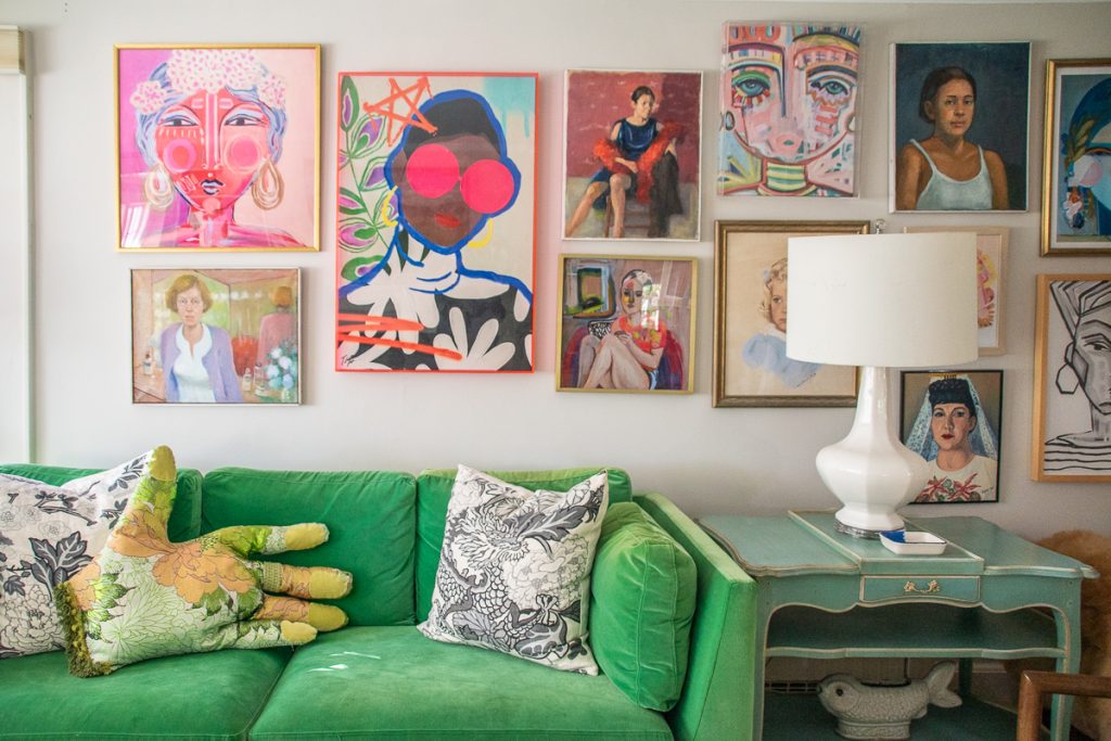 A green velvet sofa with a hand-shaped pillow sits beneath a gallery wall of colorful, eclectic portraits and paintings.