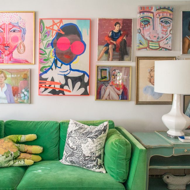 A green velvet sofa with a hand-shaped pillow sits beneath a gallery wall of colorful, eclectic portraits and paintings.