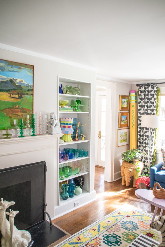 A living room featuring a fireplace mantel with green glass candlesticks, a tall shelving unit filled with colorful glassware, and a large decorative pencil standing in the corner.