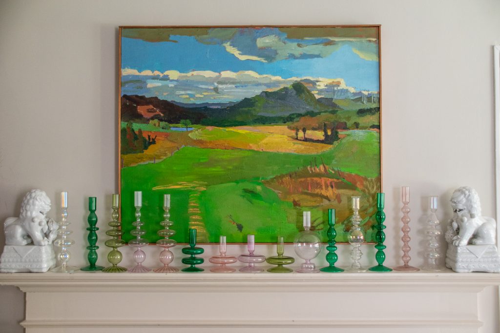 A landscape painting hanging above a mantel lined with an assortment of colorful glass candle holders and two white lion statues at each end.