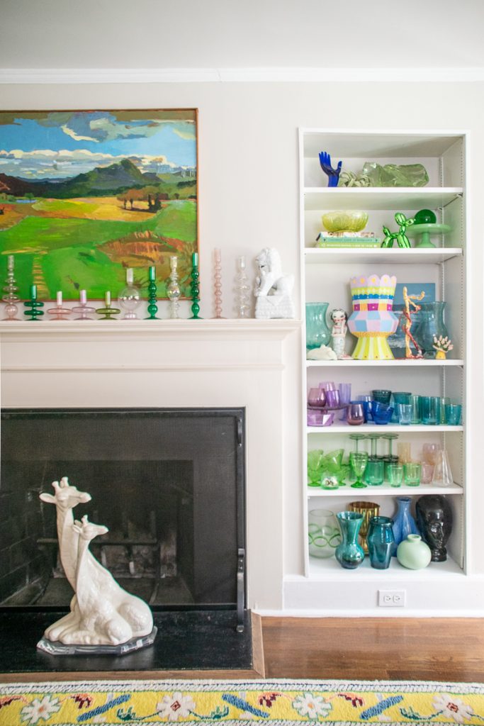 A fireplace mantle topped with a landscape painting and colorful glass candlesticks, next to a built-in bookshelf filled with glassware and decorative objects, with a ceramic giraffe sculpture in front of the fireplace.