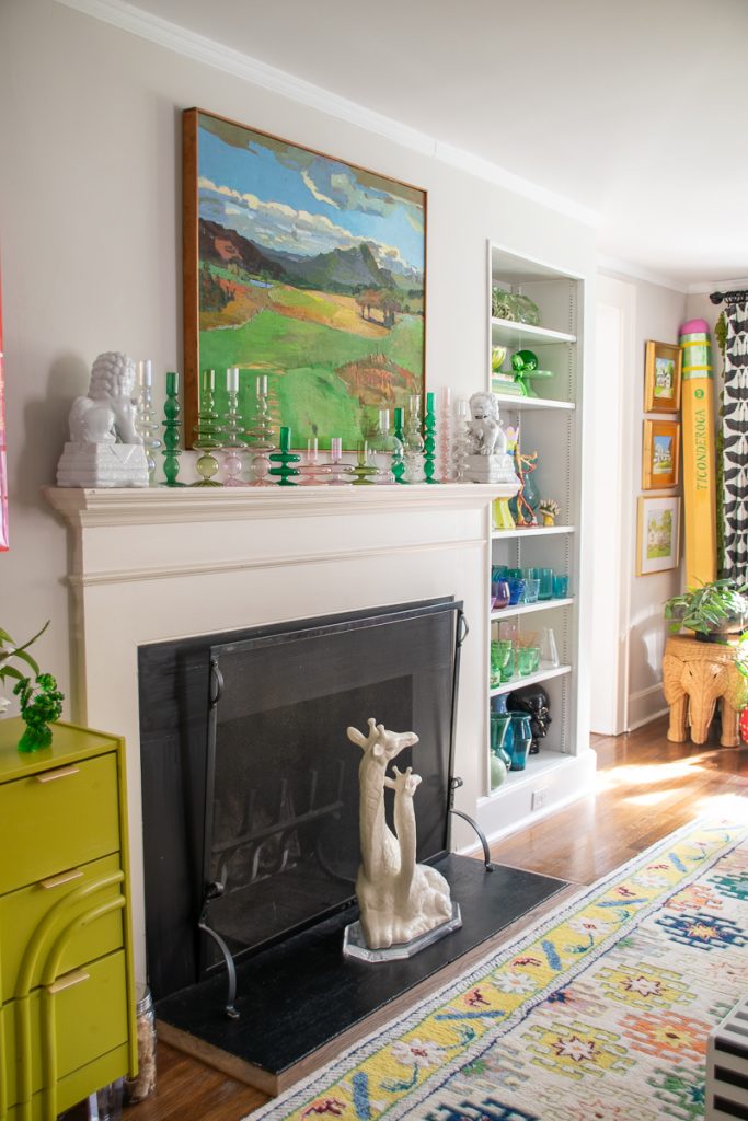 A fireplace mantel decorated with a row of colorful glass candle holders and two ceramic lion statues, with a landscape painting above it and a large decorative pencil in the corner.