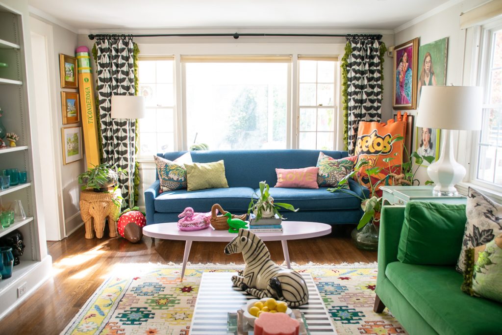A vibrant living room featuring a blue sofa, a pink oval coffee table, a striped ottoman with a decorative zebra, and eclectic decor including a large pencil and a Cheetos pillow.