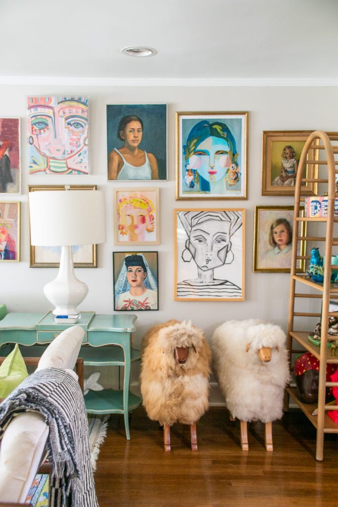 A gallery wall featuring a variety of framed portraits and abstract paintings hangs above a teal side table, a white lamp, and two fuzzy sheep-shaped footstools.