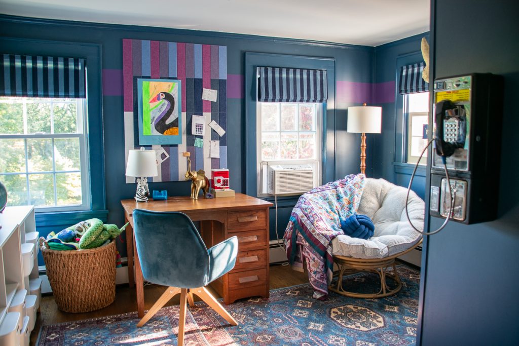 A home office with a wooden desk, blue chair, and a patterned papasan chair, featuring a wall-mounted payphone and a colorful bulletin board.